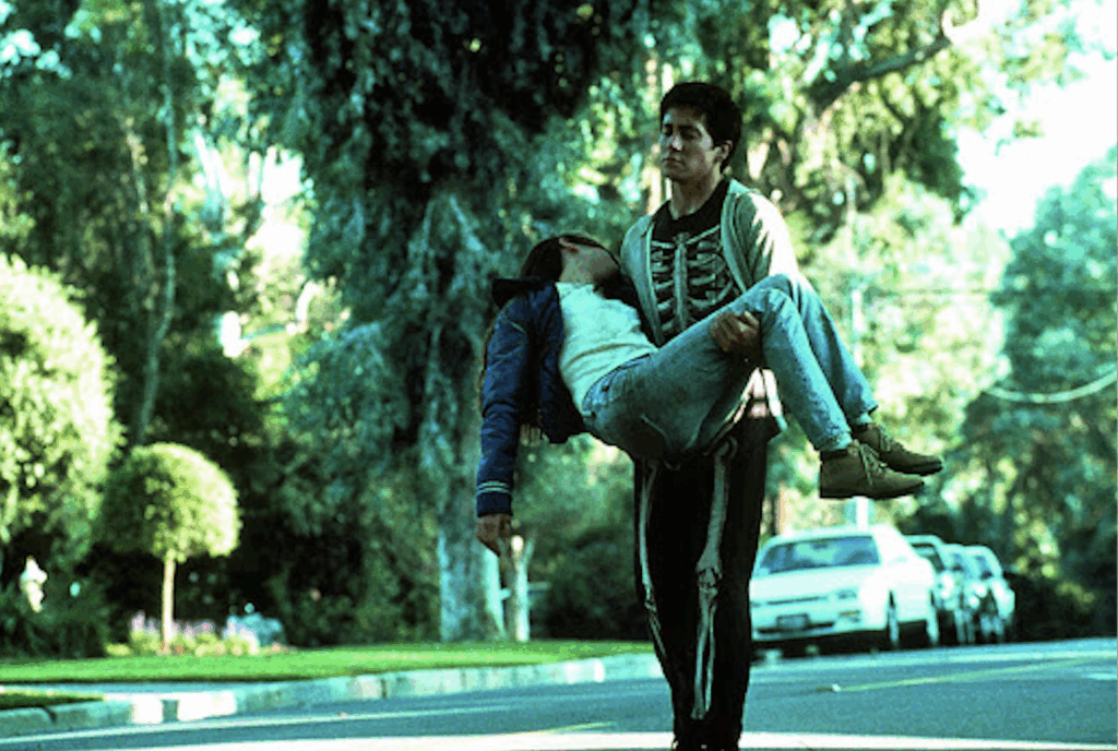a young man dressed in a skeleton onesie is carrying a girl who is slumped over in his arms. he is walking through a suburban neighborhood.
